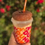 Hand holding iced coffee from Dunkin' in front of bushes with a donut placed on top of the lid.