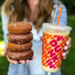 Young woman holding out four stacked donuts and an iced coffee from Dunkin' in front of her.