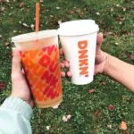 Two people holding an iced and hot Dunkin' coffee together in front of grass.